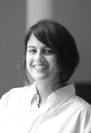 Black and white portait of Mexican author Brenda Lozano standing by a window, smiling.